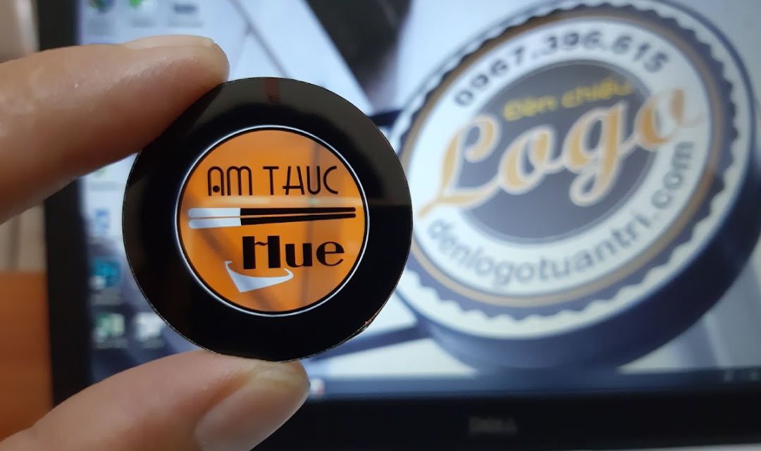 Bàn tay cầm một lăng kính tròn in logo Ẩm Thực Huế nền cam, phía sau là màn hình máy tính hiện logo Đèn chiếu Logo